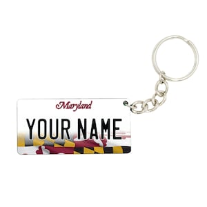 May include: A white and silver keychain with a Maryland license plate design. The plate features the Maryland state flag and the text "Maryland" and "YOUR NAME".