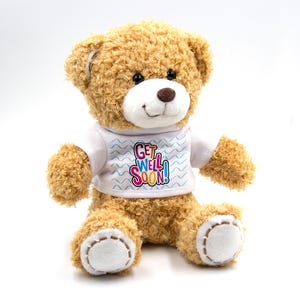 May include: A plush teddy bear with light brown fur, a white t-shirt with the text "Get Well Soon!" in colorful letters, and white accents on the paws. The bear has a friendly expression with a brown nose and black eyes.