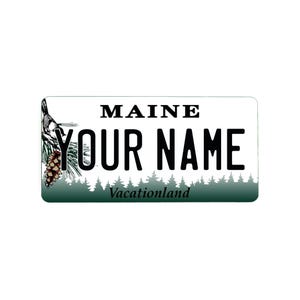 May include: A white and green personalized license plate with the text "MAINE" and "YOUR NAME" in black. The bottom of the plate has the text "Vacationland" in black. The plate has a pine tree and pine cone design on the left side.