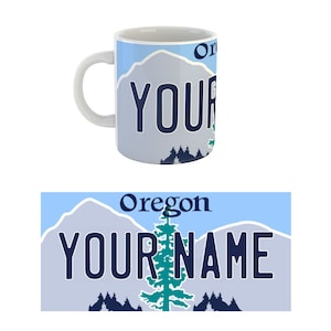 May include: White ceramic mug with a blue and white design of a mountain range and a pine tree. The mug has the text "Oregon" and "YOUR NAME" printed on it.