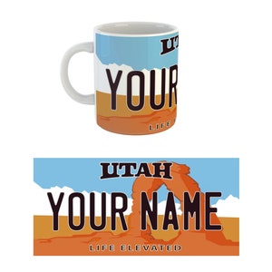 May include: White ceramic mug with a colorful design featuring a Utah license plate with the text "UTAH", "YOUR NAME", and "LIFE ELEVATED". The design also includes a depiction of Delicate Arch in Arches National Park.