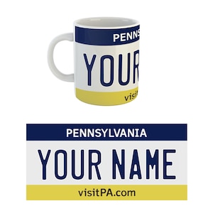 Pode incluir: Caneca de cerâmica branca com um design azul e amarelo. A caneca apresenta uma placa de matrícula da Pensilvânia com o texto "PENNSYLVANIA" e "visitPA.com" e as palavras "YOUR NAME" em letras grandes.