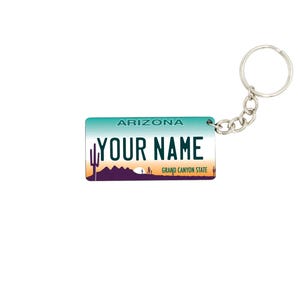 May include: A keychain featuring an Arizona license plate design. The plate is teal and white with the text "ARIZONA" and "YOUR NAME" in black. A desert landscape graphic is at the bottom. The keychain has a silver metal ring.