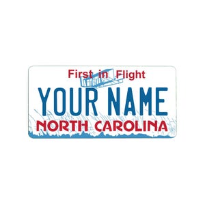 May include: A rectangular North Carolina vehicle registration plate with a white background and blue and red lettering. The top text reads First in Flight above a blue aircraft illustration. The centre shows YOUR NAME in blue, with NORTH CAROLINA below.