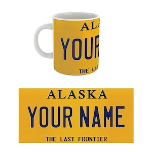 May include: Yellow mug with a blue and white Alaska license plate design. The plate reads "Alaska, Your Name, The Last Frontier".