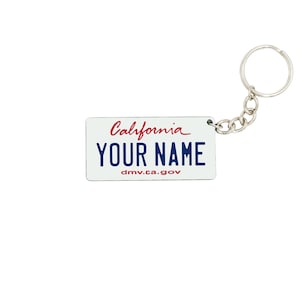 Puede incluir: Un llavero rectangular con un diseño de placa de matrícula de California. La placa dice "California" en escritura roja sobre "YOUR NAME" en letras azules. El sitio web "dmv.ca.gov" está impreso en la parte inferior. Un llavero plateado está adjunto.