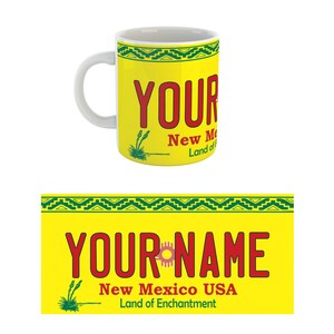May include: Yellow ceramic mug with a red and green design. The mug features a stylized license plate with the text "YOUR NAME" in red, "New Mexico USA" in black, and "Land of Enchantment" in black.