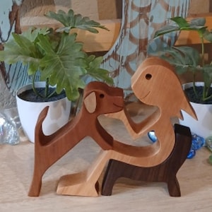 May include: Wooden sculpture depicting a person and a dog. The person is light brown, the dog is dark brown, and the bench is dark brown. The sculpture is on a wooden surface with plants in white pots in the background.
