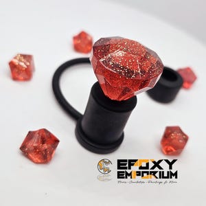 May include: A faceted, red gem-shaped bottle stopper with gold glitter accents. The stopper is attached to a black rubber ring and sits atop a black base. Several smaller gem-shaped pieces are scattered around. The logo for Epoxy Emporium is in the bottom right.