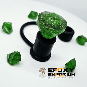 May include: A green, faceted gem-shaped bottle stopper with glitter accents, attached to a black rubber and plastic base. Several loose green gems are scattered around the stopper. The logo for Epoxy Emporium is in the lower right corner.