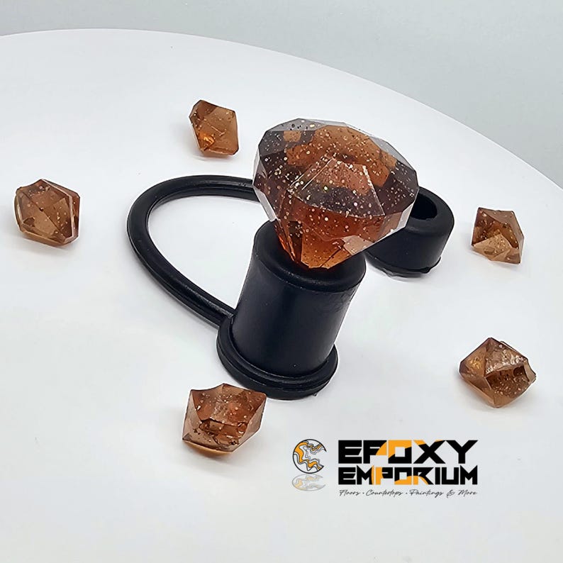 May include: A decorative bottle stopper featuring a large, amber-coloured, faceted gem design. The stopper is black with a curved handle. Several smaller, amber-coloured gem-shaped objects are scattered around. The Epoxy Emporium logo is in the lower right corner.