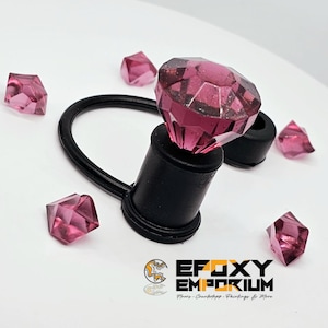 May include: A black bottle stopper with a large, faceted, pink gem on top. Several smaller pink gems are scattered around the stopper. The logo for Epoxy Emporium is visible at the bottom.