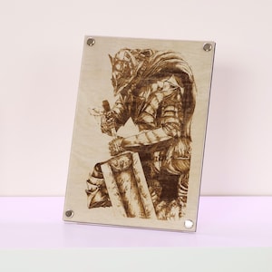May include: A wood-burned artwork showing a knight in detailed armour, kneeling and holding a sword. The artwork is mounted on a rectangular wooden panel and secured with silver screws. The knight's armour and shield are intricately detailed.