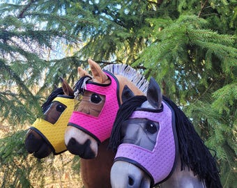 Könnte beinhalten: Drei Plüschpferde-Köpfe mit verschiedenen farbigen Fliegenmasken. Die Masken sind rosa, gelb und lila. Die Pferde sind braun, grau und weiß.