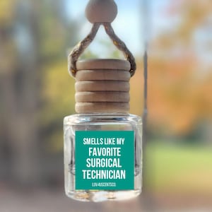 May include: A clear glass car air freshener with a wooden cap and a teal label. The label reads "Smells Like My Favorite Surgical Technician" in white text. The air freshener is hanging from a string.