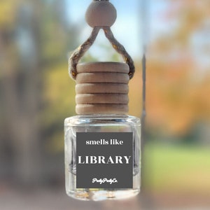 May include: A small, clear glass car air freshener bottle with a wooden cap and a jute hanging cord. The bottle features a dark gray square label with the white text "smells like LIBRARY" and "PrettyPrettyCo" printed at the bottom of the label.