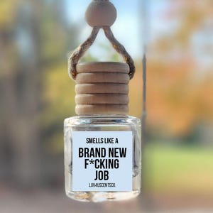 May include: A clear glass car air freshener with a wooden cap and a string for hanging. The label reads "Smells like a Brand New F*cking Job" in black text. The background is blurred with fall colors.