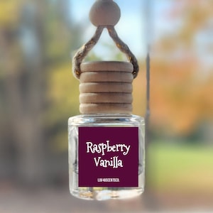 May include: A small, clear glass car air freshener bottle with a wooden cap and a jute hanging cord. The bottle features a square label with a dark purple background, white text reading Raspberry Vanilla, and LUV 4SCENTS CO. at the bottom.