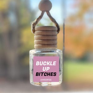 May include: A small clear glass car air freshener bottle with a light wooden cap and a jute hanging cord. The bottle features a pink square label with the white and black text BUCKLE UP BITCHES and the smaller text LUV 4USCENTSCO printed below.