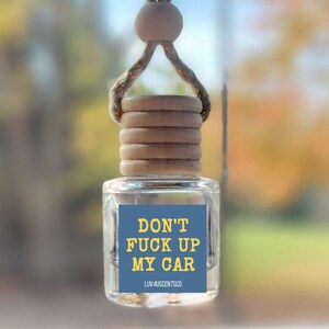May include: A small, clear glass car air freshener bottle hanging from a rearview mirror by a tan twine cord. The bottle features a wooden cap and a blue square label with yellow text that reads DON'T FUCK UP MY CAR and LUV4SCENTSCO.