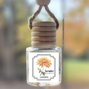 May include: A car air freshener with a wooden bead and string hanger. The bottle is clear glass with a label that says "November" and features a yellow chrysanthemum flower. The label also says "Luna Aromatics".