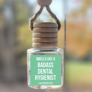 May include: A clear glass car air freshener with a wooden cap and a hemp string for hanging. The label reads "Smells Like A Badass Dental Hygienist". The background is blurred, showing a glimpse of trees.