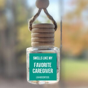 May include: A clear glass car air freshener with a wooden cap and a teal label that reads "Smells Like My Favorite Caregiver" and "Luv4uscents.co". The air freshener hangs from a string with a wooden bead.