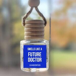 May include: A clear glass car air freshener with a blue label that reads "Smells Like a Future Doctor." The air freshener is suspended by a natural rope and wooden bead. The bottom has a wooden cap.
