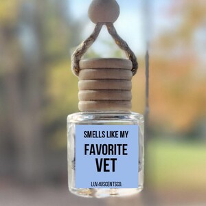 May include: Clear glass car air freshener with a wooden cap and a light blue label. The label reads "Smells Like My Favorite Vet" in black text. The air freshener hangs from a string and wooden bead.
