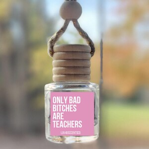 May include: A clear glass car air freshener with a pink label that reads "ONLY BAD BITCHES ARE TEACHERS". The air freshener is suspended by a natural rope and wooden bead. The top is made of stacked wooden discs.