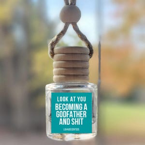 May include: Clear glass car air freshener with a wooden cap and a teal label. The label reads "LOOK AT YOU BECOMING A GODFATHER AND SHIT". The air freshener hangs from a string and wooden bead.