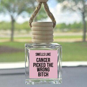 May include: Clear glass car air freshener with a wooden top and a pink label. The label reads "Smells Like Cancer Picked The Wrong Bitch" in black text. The air freshener hangs from a gold and beige rope.