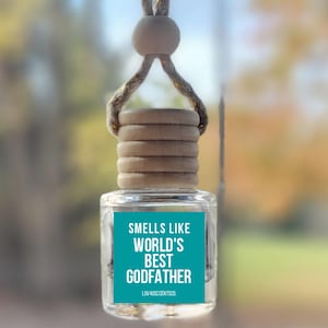 May include: A clear glass car air freshener with a wooden cap and a teal label. The label reads "Smells Like World's Best Godfather". The air freshener hangs from a string with a wooden bead.