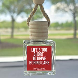 May include: A clear glass car air freshener with a wooden top and a red label. The label reads "LIFE'S TOO SHORT TO DRIVE BORING CARS" in white text. The air freshener hangs from a gold-threaded rope.