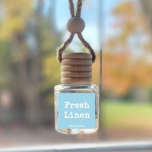 May include: A car air freshener with a light blue label that says "Fresh Linen" and a brown wooden cap. The air freshener is hanging from a rearview mirror in a car.