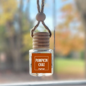 May include: A car air freshener with a brown wooden cap and a clear glass bottle. The bottle has a label that says "Pumpkin Chai" with a small logo underneath. The air freshener is hanging from the rearview mirror of a car.