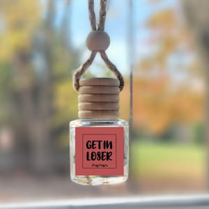 May include: A pink car air freshener with a wooden top and a white string. The air freshener has a square label with the text "GET IN LOSER" in black.