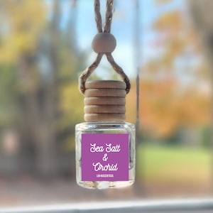 May include: A car air freshener hanging from the rearview mirror. The bottle is clear and contains a purple liquid. The label reads "Sea Salt & Orchid" with the text "LILA AROMA DIFFUSER" below.
