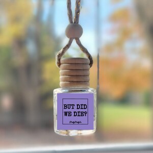 May include: Purple car air freshener with a white string hanging from the rearview mirror. The air freshener has a white label with black text that reads "But did we die?" and the text "Pretty Pretty Co."