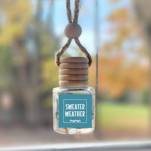 May include: A blue car air freshener with a wooden cap and a string for hanging. The label reads "Sweater Weather" with the text "papyrus" underneath.