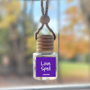 May include: A purple car air freshener with the text "Love Spell" and the words "LILA ESSENTIALS" in smaller font. The air freshener is hanging from a rearview mirror with a brown rope and wooden beads.