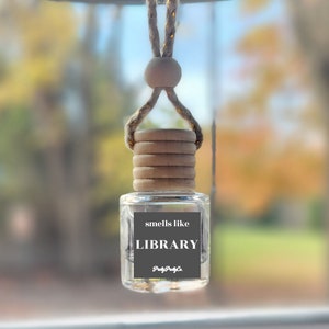 May include: A clear glass bottle with a wooden cap and a gold string hanging from the rearview mirror of a car. The bottle has a label that reads "smells like LIBRARY" with a small logo that says "Pretty Puff Co."