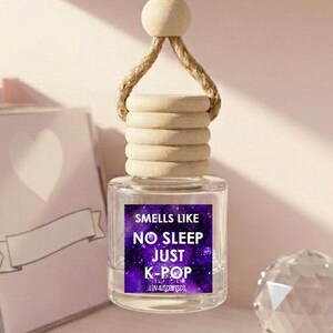 May include: A clear glass car air freshener bottle with a wooden cap and a thick jute hanging cord. The label features a purple galaxy background with the white text: SMELLS LIKE NO SLEEP JUST K-POP. The bottle is suspended by a natural fiber rope.