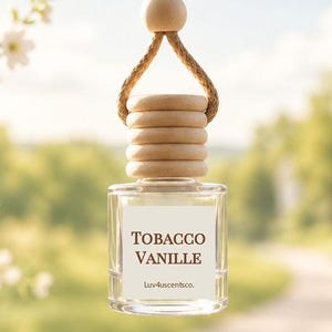 May include: A clear glass car air freshener bottle with a light wood cap and a tan rope hanger. The square bottle features a white label with the brown text Tobacco Vanille and Luv4uscentsco. printed on the front, suspended by a wooden bead.