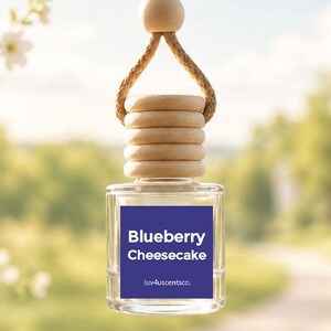 May include: A small, clear glass hanging air freshener bottle with a light wood cap and a matching wooden bead on a tan cord. The square label is dark purple with white text that reads Blueberry Cheesecake and luv4uscentsco. at the bottom.