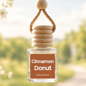 May include: A small, clear glass car air freshener bottle with a light brown wooden cap and a matching wooden bead on a tan hanging cord. The bottle features a square label with the text Cinnamon Donut and luv4uscentsco. in white font.