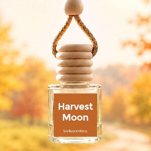 May include: A clear glass car air freshener bottle with a wooden cap and a braided hanging cord. The square bottle features a burnt orange label with the white text Harvest Moon and the smaller text luv4uscentsco. centered on the front.