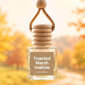 May include: A small, clear glass car air freshener bottle with a light brown wooden cap and a braided cord for hanging. The square bottle features a tan label with the white text Toasted Marshmallow and the brand name luv4uscentsco. printed below.
