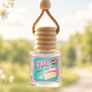 May include: A small, clear glass bottle air freshener hanging from a tan rope. The bottle features a wooden cap and a label with a pink cake illustration, the text CAKE Bakery shop, and a small red seal. The design is square with rounded corners.