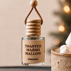 May include: A hanging car air freshener with a clear glass bottle, a light wooden cap, and a tan label featuring the text TOASTED MARSHMALLOW and Luv4uscentsco. The bottle is suspended by a tan cord with a small wooden bead accent.